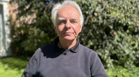 Gary Widdowfield is sitting on a garden chair. He is looking directly at the camera. he has grey hair and is wearing a navy jumper with a black top underneath.