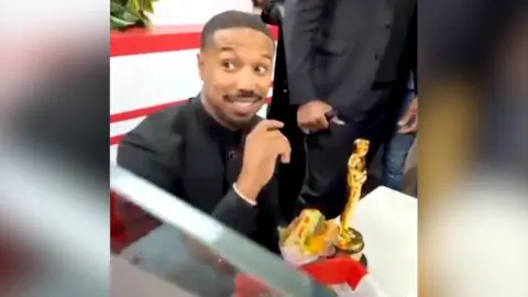 Michael B Jordan smiles at an In-N-Out Burger restaurant as he holds a burger. His Oscar sits on the table next to him.