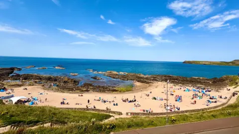 BBC Weather Watcher AlicelouiseP A wide landscape picture of a beach crowed with people. The sea and the sky are blue and there are only a few whispy clouds.