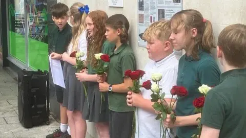 Some of the pupils involved in the creation of the plaque are standing by the wall it is displayed on, holding red and white roses.