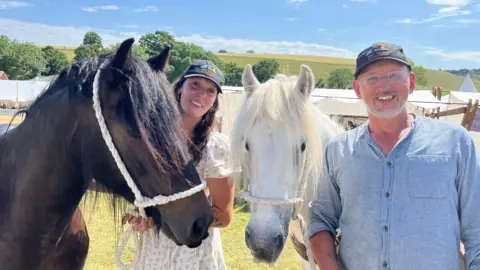 Two fell ponies - one brown and one white - pictured with their handlers, one man and one women. It is a sunny day with the light shining brightly on the grass and fields in the background. There are a few white tents behind them.