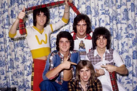 Getty Images The Bay City Rollers pictured in the 1970s. They are five young men with spikey and shoulder-length hair, wearing shirts incorporating tartan. One of them is holding up a tartan scarf. They are standing against a blue and white Willow pattern curtain 