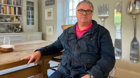 Stephen sits in a kitchen looking at the camera. His left hand is on the kitchen table which is next to him. He's wearing a dark blue denim jacket with a pink, collared t-shire under it. Behind him is a cabinet with books to his left and to his right there are large decorative cultarly hanging from the wall.