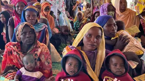 Women in colorful headscarves sitting in a chaise holding some children on their laps in a makeshift reception area at the Adre border post in Chad