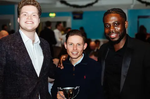 Bedford Borough Council Three young men look directly at the camera and smile. The man on the right has his right arm on the right shoulder of the man in the centre, who is holding a trophy.