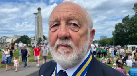 A person wearing a suit jacket with medals and a tie stands in the foreground on Plymouth Hoe, with a large crowd gathered behind them. In the background, people are walking and sitting on the grass near a tall naval memorial column, with more event activity taking place across the waterfront.
