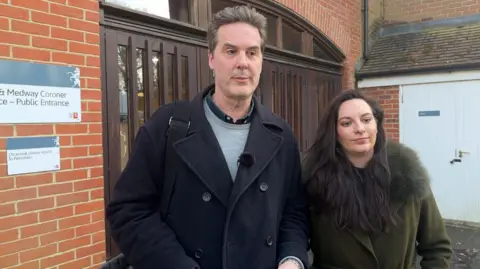 Kent Mathews wearing a dark jacket, grey jumper and navy shirt stands outside Kent & Medway Coroner's Court next to solicitor Frankie Rhodes wearing a khaki jacket with matching fur trim. 