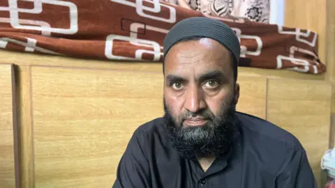 Man with beard dressed in black and wearing a black cap looks towards camera - behind him is what looks like a bunk bed.