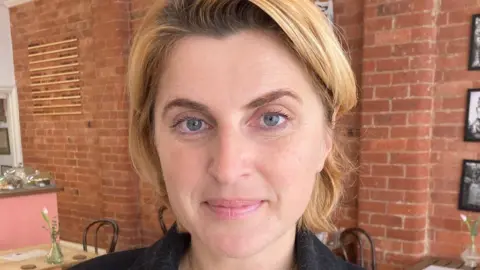 Solomiia Blagitko, standing in a room, looking at the camera and smiling. She has blonde hair, to her chin and a black top on. There are tables and chairs behind her. 