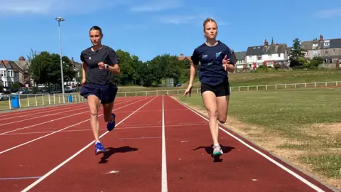 Jennie Dennett/BBC Honor and Gretta running on the track side-by-side. They are both mid-stride and Honor is on the left with blue trainers and Gretta on the right with grey trainers. They are both wearing shorts. Honor's are blue and floaty and Gretta's are black and tight.