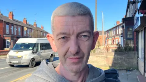 Richard poses for the camera - along the pavement of the proposed cycle lane. He has shaven, grey hair. He is wearing a grey hoodie and there is a white van and a row of red brick houses over his right shoulder. 
