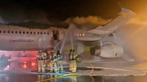 Reuters Six firefighters on an airport runway spray hoses onto the side of a jet airplane at night