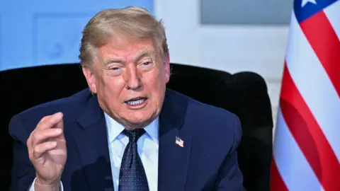 US President Donald Trump speaks while sitting down at the G7 summit, with an American flag next to him