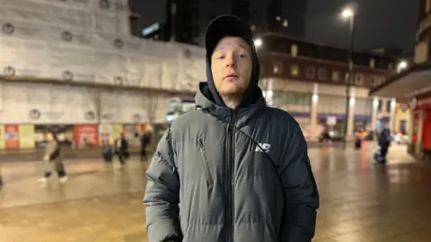 Gareth stands in Leeds city centre, wearing a black cap and black coat. He has his hands in his pockets