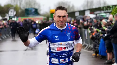PA Media Kevin Sinfield waves a gloved hand to the camera while running. He is in a blue T-shirt with a blue running vest over the top. It has the number seven emblazoned on it alongside the names of other sponsors. Behind him crowds of people behind barriers are clapping.