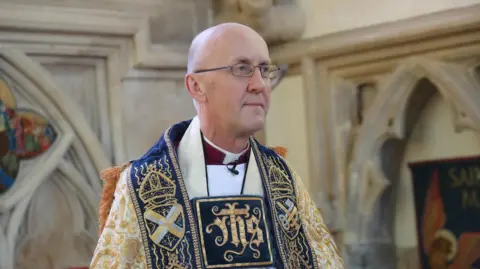 Jason Bryant Bishop Michael Beasley is pictured in his ceremonial robes looking off to the right. He is standing in a church, with arches behind him. 