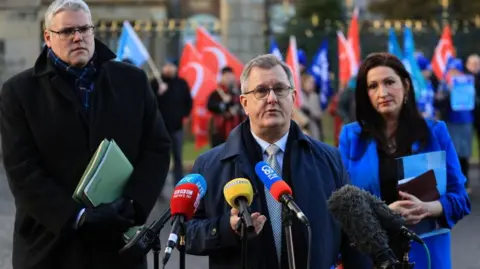 PA Media Two men and a woman stand side by side in front of microphones. the man on the left is tall with glasses. he is wearing a black overcoat and scarf. The man in the middle is speaking. He has grey hair and round glasses. He is wearing an overcoat, gold tie and white shirt. The woman has long dark hair and is wearing a blue coat. There are blue and red flags being held by a blurred crowd behind them.
