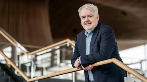 Carwyn Jones leaning on a bannister inside the Senedd. He is wearing a dark suit with a checked blue shirt, and has grey hair and a short beard.