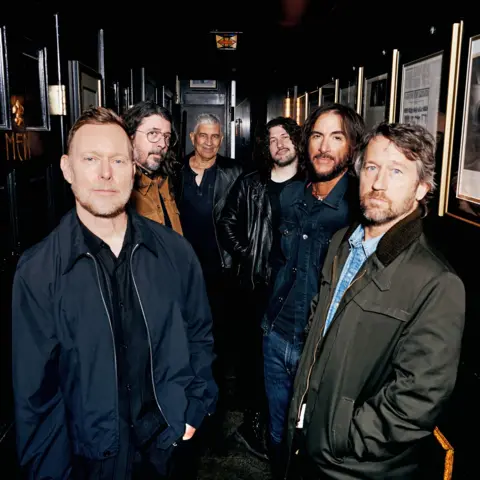 Elizabeth Miranda The six members of Foo Fighters stand side by side in a long, wood-panelled corridor. They're looking directly at the camera.