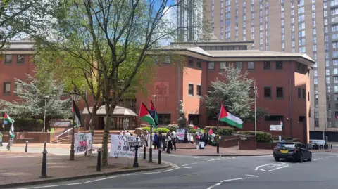 A group of people, flying the flag of Palestine outside a large red brick court building