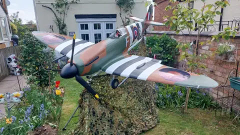 Ray Burden A large model Spitfire plane in a garden. The plane is dark green, white, black and brown with black propellers on its nose. It has an RAF circular logo on the side and camouflage netting underneath it.