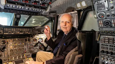 PA Media John Britton sat in the cock pit of Concorde. He is holding the new 50p coin up to the camera.