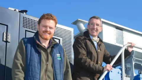 Andrew Muir with Jack Blakiston Houston from BH Estates. Andrew Muir is wearing a brown coat, light blue shirt, yellow bow tie and glasses, and has his hands together. Jack Blakiston Houston has ginger hair and beard, wearing a blue puffer vest and green jumper. They both stand on a platform with farm equipment in the background.