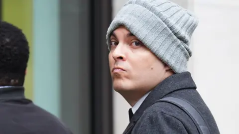 Che Homersham entering Southwark Crown Court wearing a charcoal jacket, grey beanie and with a black rucksack over one shoulder.