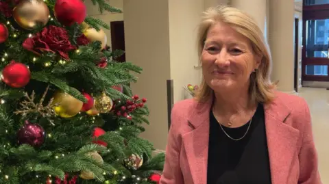 BBC A woman wearing a pink blazer stood near Christmas tree in a hallway. She is smiling.