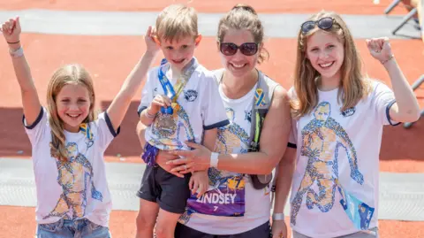 PA Media Lindsey wears running shorts and sunglasses and has her hair in a ponytail. She is holding her son in her arm. Either side of her are girls who look about 10 and 13, all cheering. They all have matching white T-shirts with Rob on.
