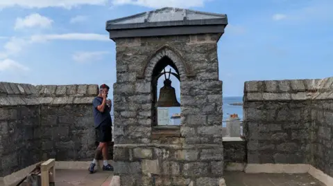 Manx National Heritage The Church bell, in the background is the sea, and stood next to it is a man on the phone. 