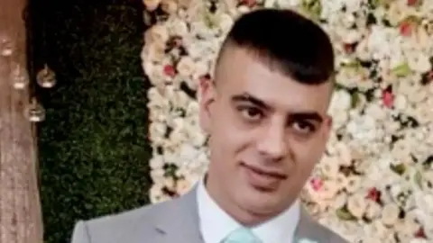 West Yorkshire Police A man in his thirties with dark hair, stood against a flowery backdrop. He is wearing a grey suit and blue tie. 