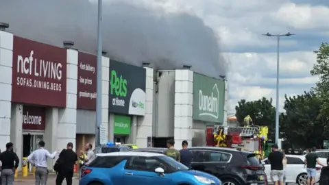Crowds of shoppers look on as emergency service workers attend a fire at a store in a retail park. Smoke can be seen billowing from the Dunelm store. 