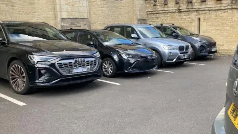 Four cars in a car park. Three are black, one is grey. There's another car to the right of the picture, just out of shot.