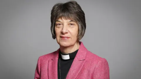 An official portrait of the Bishop of Gloucester, an older woman with a short dark grey/brown bob, a pink blazer, a black top and a vicar's dog collar. She is stood against a light grey background.
