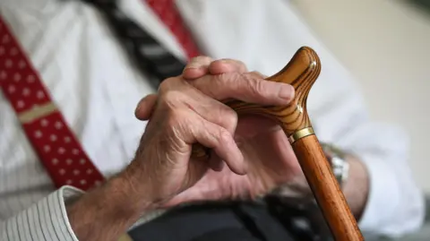PA Media A man's hands rest on the top of a walking stick. He is wearing a white striped shirt and red braces. 
