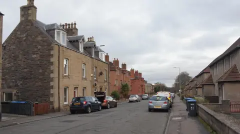 Ian S A suburban street in Galashiels with houses up both sides and cars parked on both sides of the road as well
