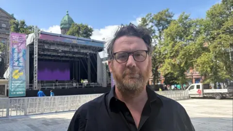Pádraig Ó Duinnín, a man with grey hair, a red and grey beard. He has glasses and is wearing a black shirt. He is standing in front of a stage at City Hall.