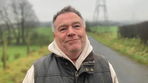 William Sloan. He has short light brown hair, wearing a khaki wax zip up vest with bream hoody underneath. Behind him is a country road with fields and an electricity pile on.