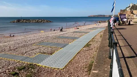 East Devon District Council Sidmouth Town Beach with track matting going across and down the beach. There are people on the beach by the water. 