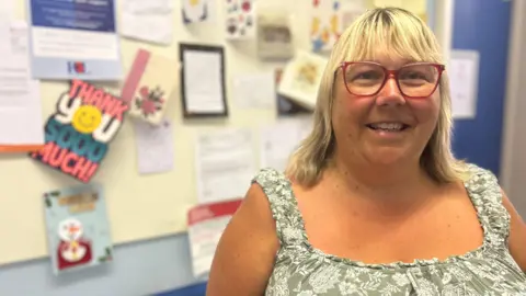 A woman with blonde shoulder length hair and fringe and red-framed glasses smiles in front of a cream wall with thank you cards pinned to it. She is wearing a green and white floral sleeveless dress.