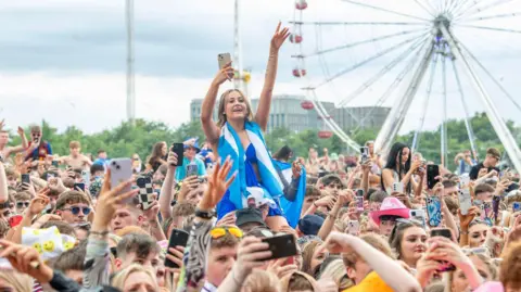PA Media A crowd of people with their hands in the air and people hoisted onto shoulders. One girl on someone's shoulders in the foreground wears all blue with a saltire flag draped around her neck. A Ferris wheel can be seen in the background.