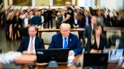 EPA Donald Trump sits in court, flanked by his lawyers, with others in court behind him. Much of the image is blurry with Trump in focus in the centre.