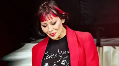 Saffron Sprackling Saffron in a red jacket with a black T-shirt with white writing on it. She has brown hair with red streaks.