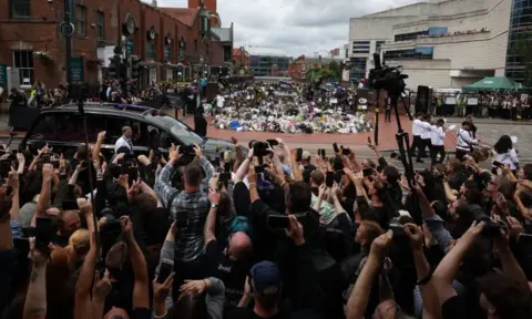 EPA A line of musicians are playing in front of a funeral car, as they pass Black Sabbath bench in Birmingham's Broad Street, which is festooned in floral tributes. In the foreground there is a sea of fans and photographers craning to get a picture.