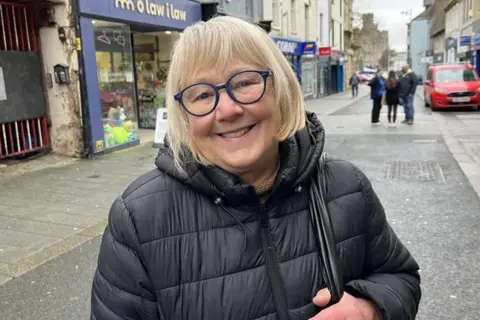 Gwenda Harding from Llanberis, Gwynedd. She is stood on the high street in a thick coat and wears glasses. she is smiling at the camera.