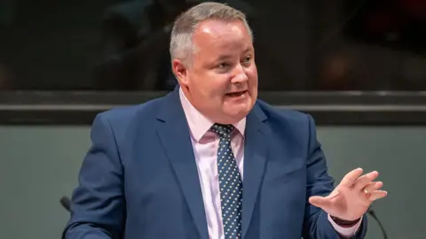 Darren Millar gesturing with one hand, wearing a dark suit, pink shirt and a blue tie