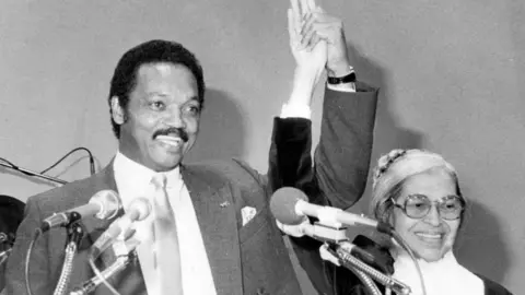 Getty Images A black and white film photo of Jesse Jackson pictured with Rosa Parks, holding hands and appearing united.