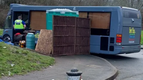 The blue vehicle is has wood against the windows on the inside. A police officer stands by the door looking in. There is various debris of wood, a plastic-looking tank, near the entrance. What looks like a mobile toilet next to a fence is set up by the the side of the vehicle.