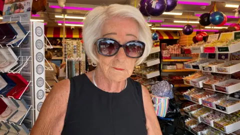 A woman in a black sleeveless dress looks at the camera. She has dark sunglasses and a silver necklace. She is standing in front of shelves full of sweets and toys.